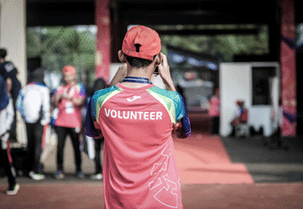 A young volunteer, dressed in a t-shirt that says 'Volunteer' prominently, photographs a volunteering event.